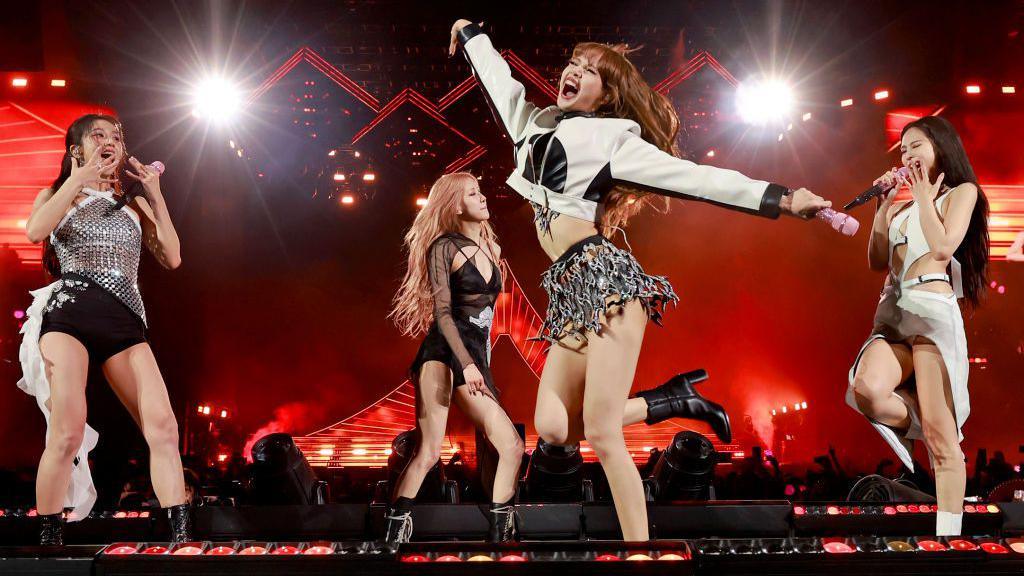 Lisa from BLACKPINK throws her arms up as she dances at the 2023 Coachella Valley Music and Arts Festival, along with other band members. They are dressed in black, white and silver on a stage lit up in red. 