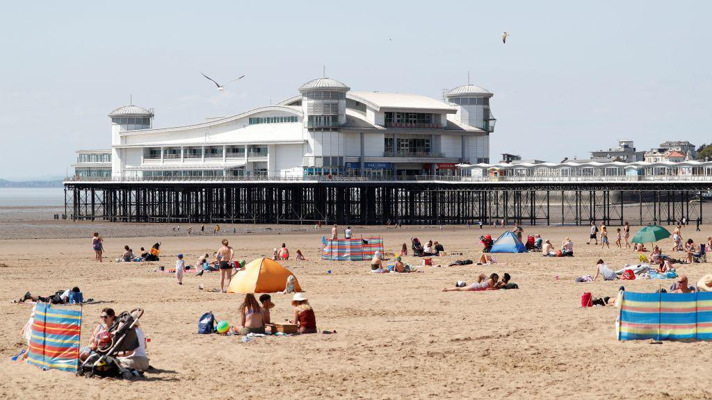 Families sitting on a beach on a sunny day with a long pier in the background