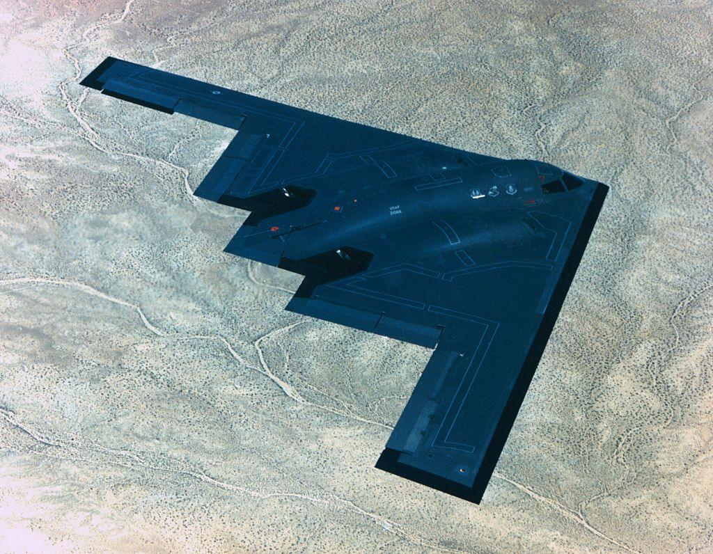 B-2 Spirit stealth bomber, painted all black, flying from left to right high above a desert landscape.
