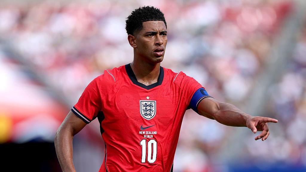 Jude Bellingham, wearing a red England shirt with a white number 10 in the centre beneath the badge, extends his left arm and points his index finger
