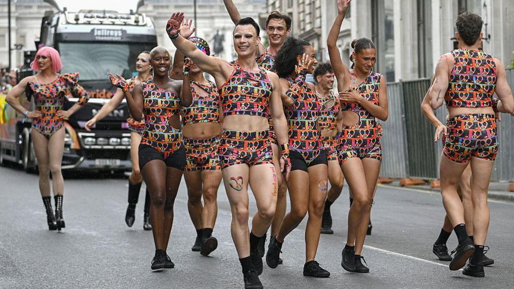 A group of men and women dressed in identical shorts and cropped tops of multi-coloured fabric wave at the crowd as they march in the parade. 