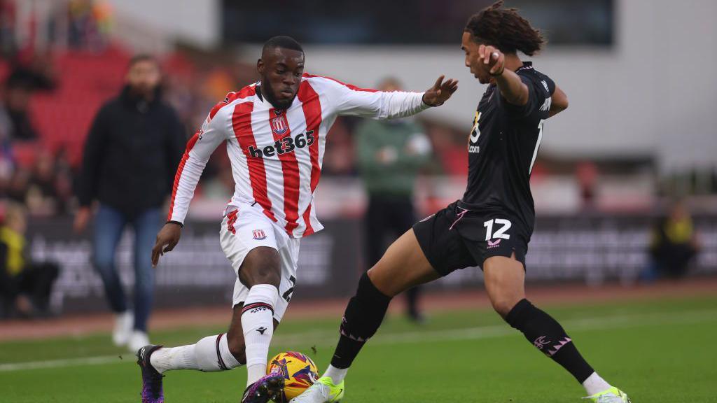 footballer in red and white stripes competes for ball with footballer in black strip