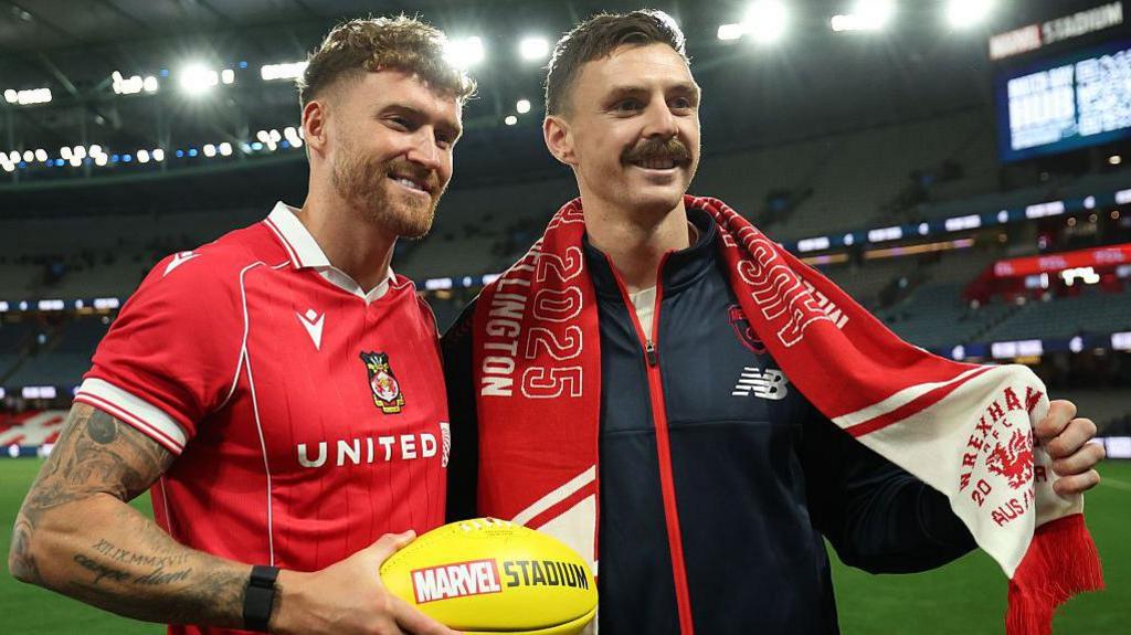 Dan Scarr holds an Aussie Rules football in a Wrexham kit, next to Melbourne Demons player Jake Lever in a navy tracksuit and a Wrexham scarf.