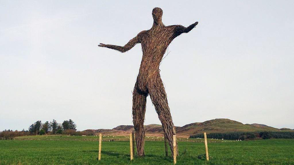 A large wicker sculpture of a man with his arms held out, standing in the middle of a grass field