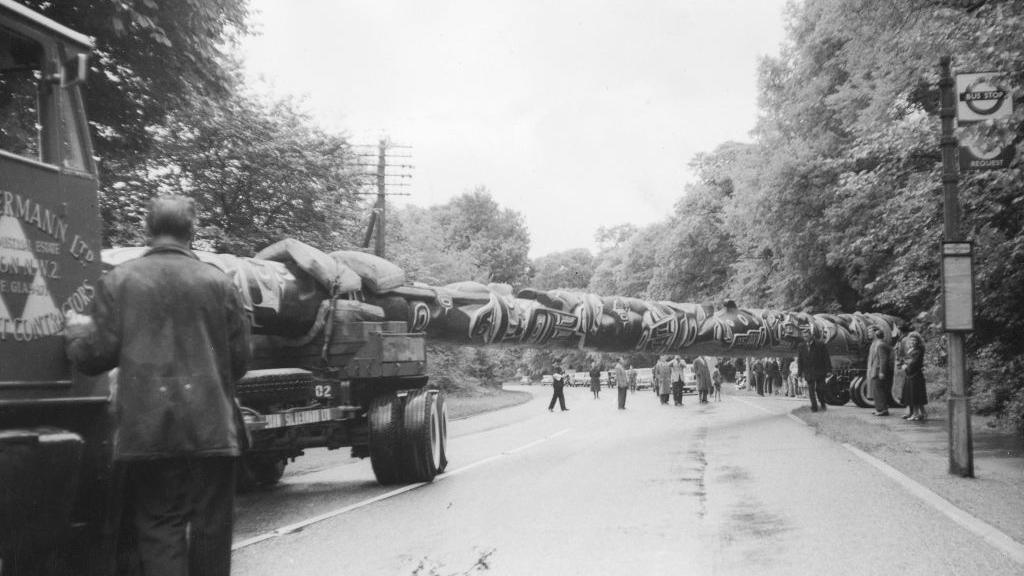 A black and white image showing the totem pole on the back of a lorry and stretching across a road