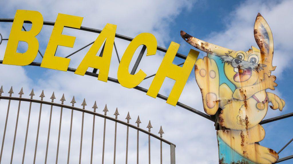 An iron gate with the word "beach" in yellow letters above and a rusty, faded sign in the shape of a rabbit.