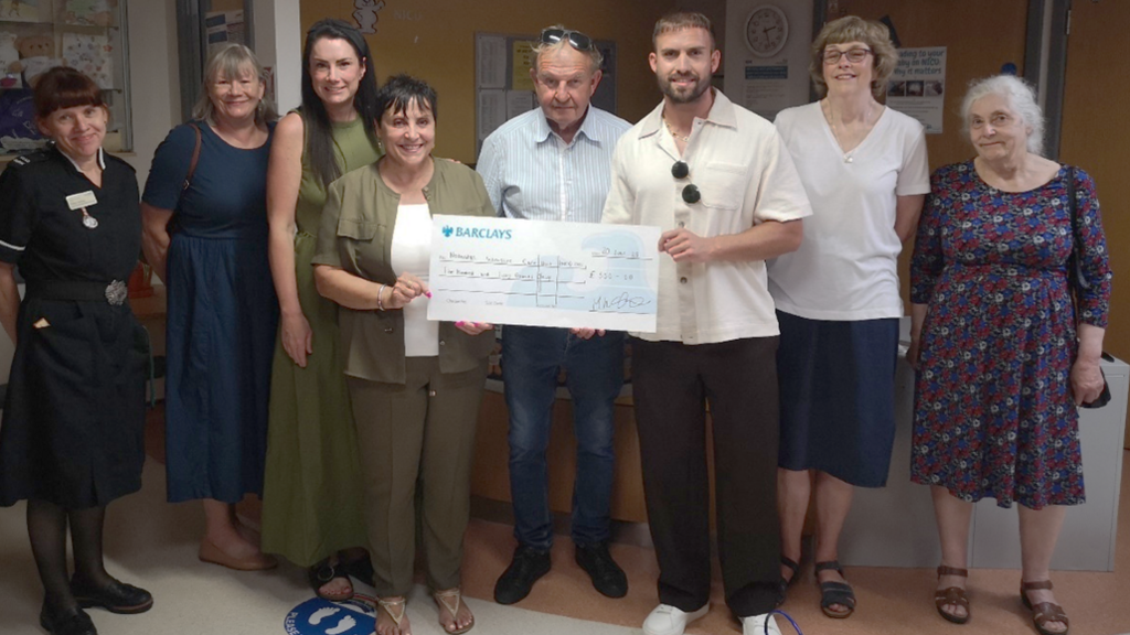 Eight people are standing in a row in a hospital ward. The three in the centre are holding a large cheque.