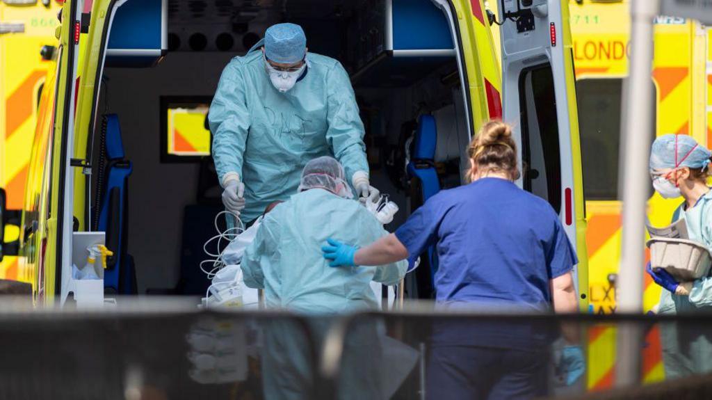 Two medical workers in full PPE - green overwear, hat and masks, helping someone in a wheelchair into the back of an ambulance. A nurse in normal scrubs and mask is helping them.
