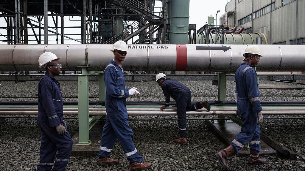 Gas workers dressed in protective gear walk past a large pipeline labelled 'natural gas'.