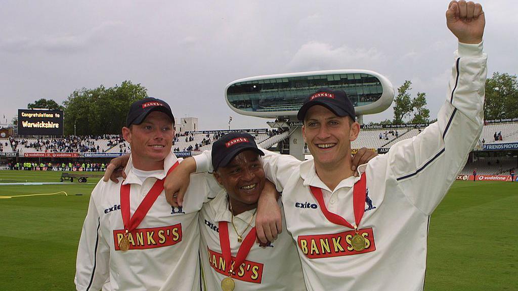 Keith Piper stands in the middle of three players with Ian Bell to his right and Jim Troughton to his left. All three are wearing gold medals on right ribbons around their necks after winning the Benson and Hedges Cup at Lord's. They are all wearing navy blue caps and Troughton has his left arm raised. The media centre at Lord's is visible behind them as is a black digital scoreboard displaying the message "Congratulations Warwickshire" in gold letters.