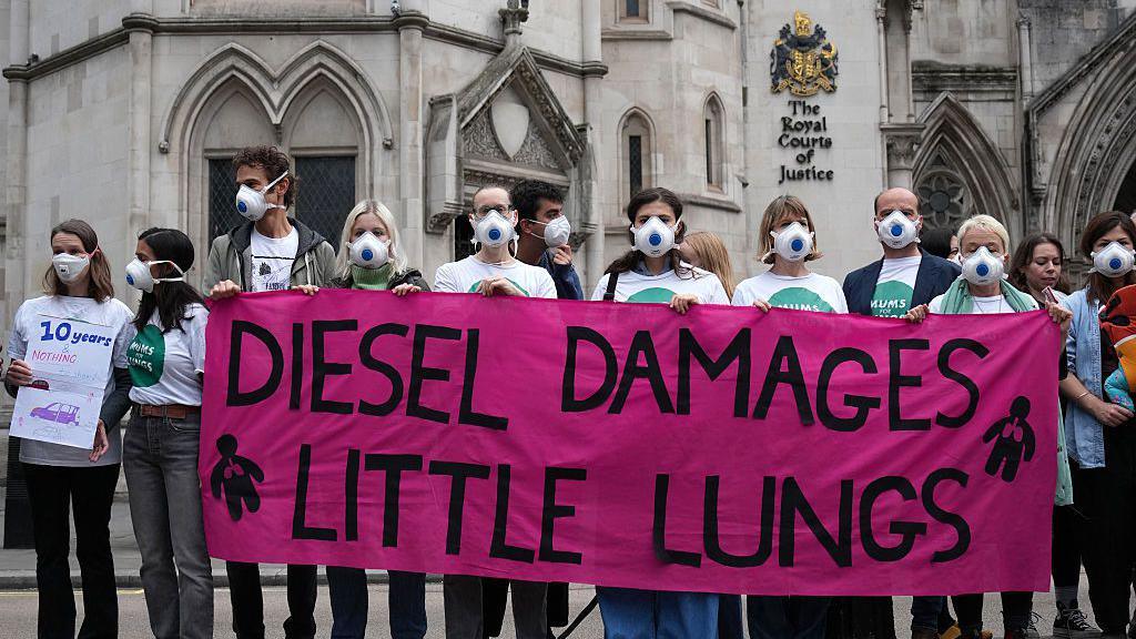 Thirteen people wearing gas masks and holding a sign that says 'diesel damages little lungs'. They are standing in front of the Royal Courts of Justice