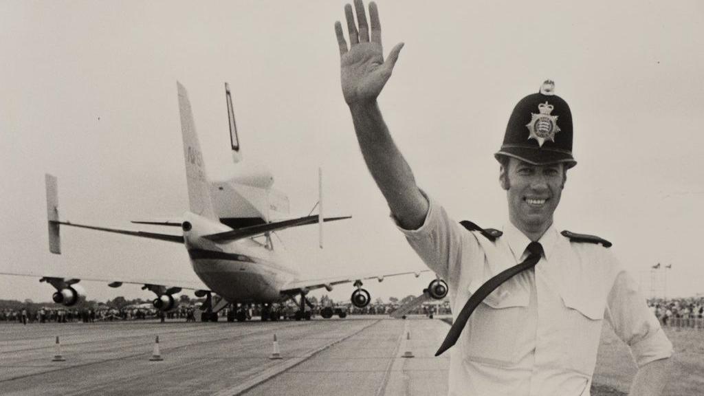 A black and white photo of officer Brian Bishop. He is pictured in police uniform at Stansted Airport. He has one arm up in the air and the other resting on his hip. He is smiling to the camera and an aeroplane is parked behind him.