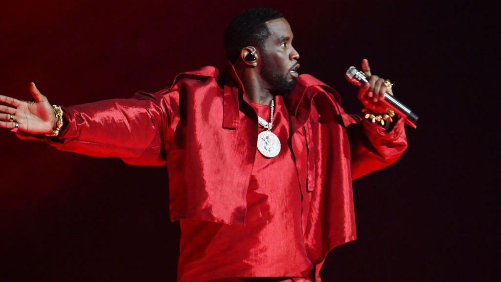 Sean "Diddy" Combs appears on stage holding a mic for a performance. He is wearing all red with a thick gold chain with a large gold medallion. 