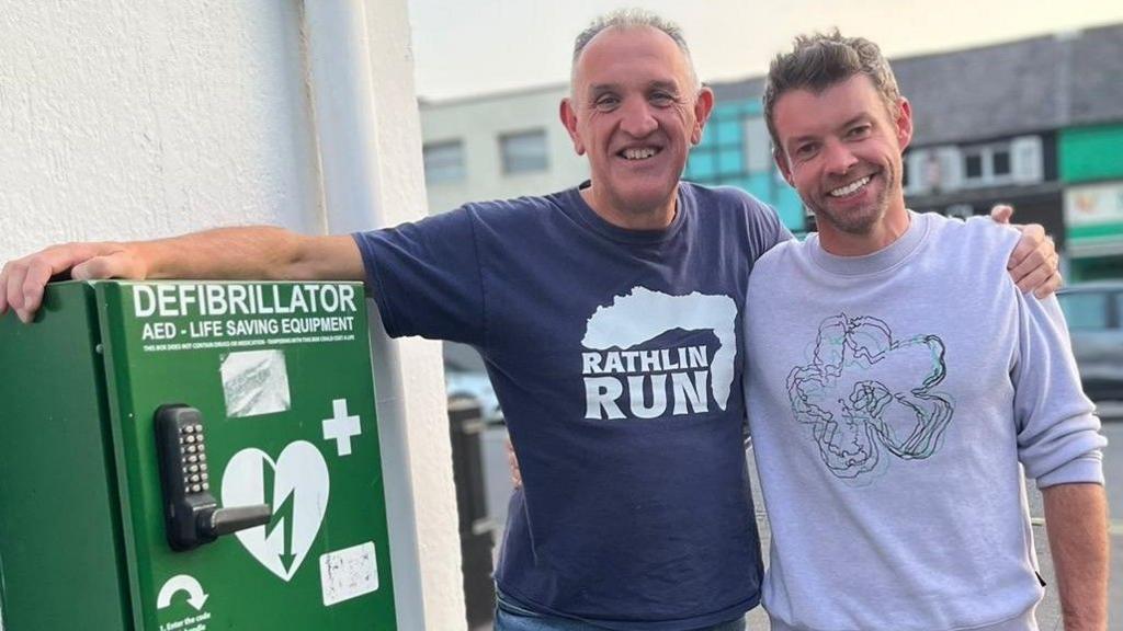 Peter O'Hare stands to the right of John Deery. They are grinning for the camera and are standing next to a defibrillator in a big green box attached to a wall. They are standing in a street. John is wearing a t-shirt and jeans, while Peter is wearing a white jumper with a logo on it. He has brown hair and is wearing a smart watch with a red strap.