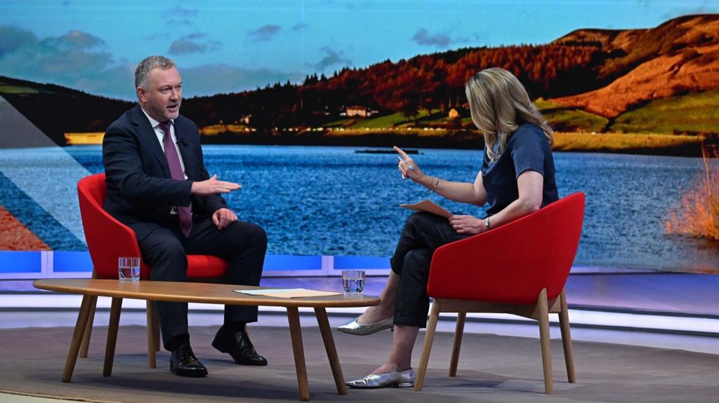 Steve Reed (L) sitting down wearing a suit and tie with his right hand partially raised as he answers questions to Laura Kuenssber (R)