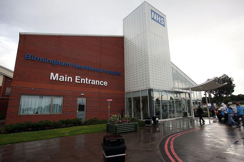 A red brick building with a chrome and glass tower in the centre. A sign says "Birmingham Heartlands Hospital" in blue on the redbrick wall. 