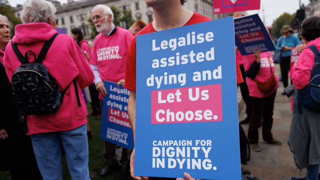 Protestors outside the Houses of Parliament. They are wearing pink sweatshirts that say "Dignity in Dying" in white letters on them.