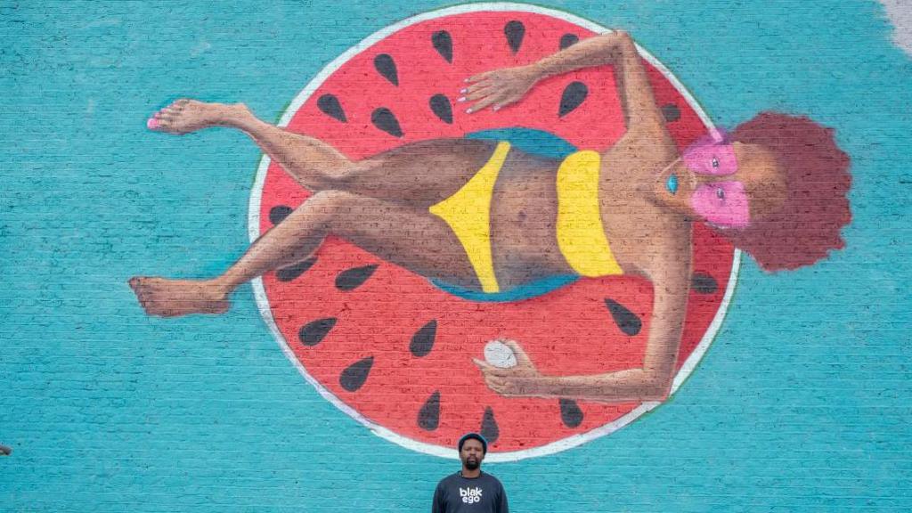 An image of South African artist Breeze Yoko standing in front of a mural of a woman wearing a yellow bathing suit, sitting in a float in the shape of a watermelon on 31 July 2024.