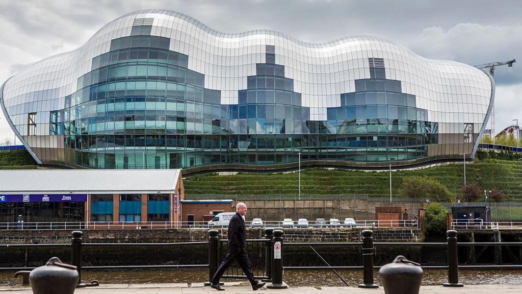 The Glasshouse International Centre for Music viewed from Newcastle Quayside. A large building, it has a futuristic shape and is made up of hundreds of glass panels. In the foreground, a man is walking along the Quayside towards to the right of the photo.