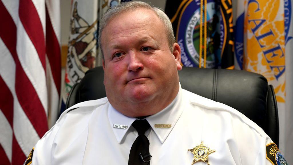 Former Culpeper County Sheriff Scott Jenkins wearing a white shirt, black tie with a gold badge on the right side