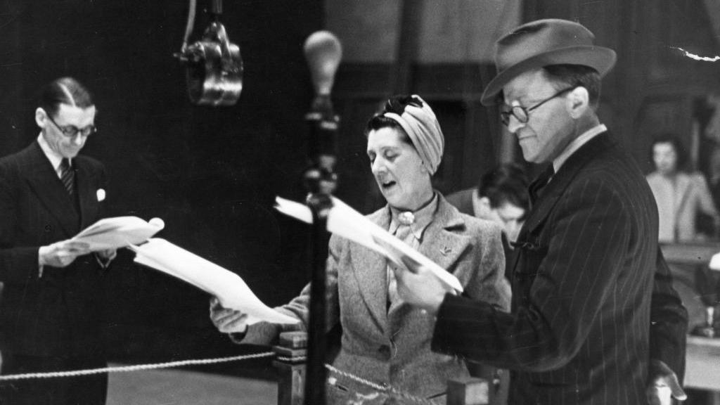 A black and white picture of a man and woman reading scripts at a microphone.