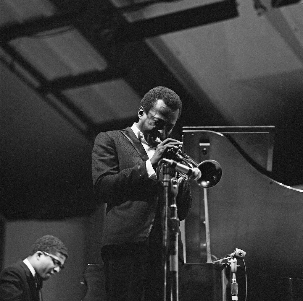 A black and white photo of Miles Davis playing the trumpet with Herbie Hancock in the corner playing keyboards.