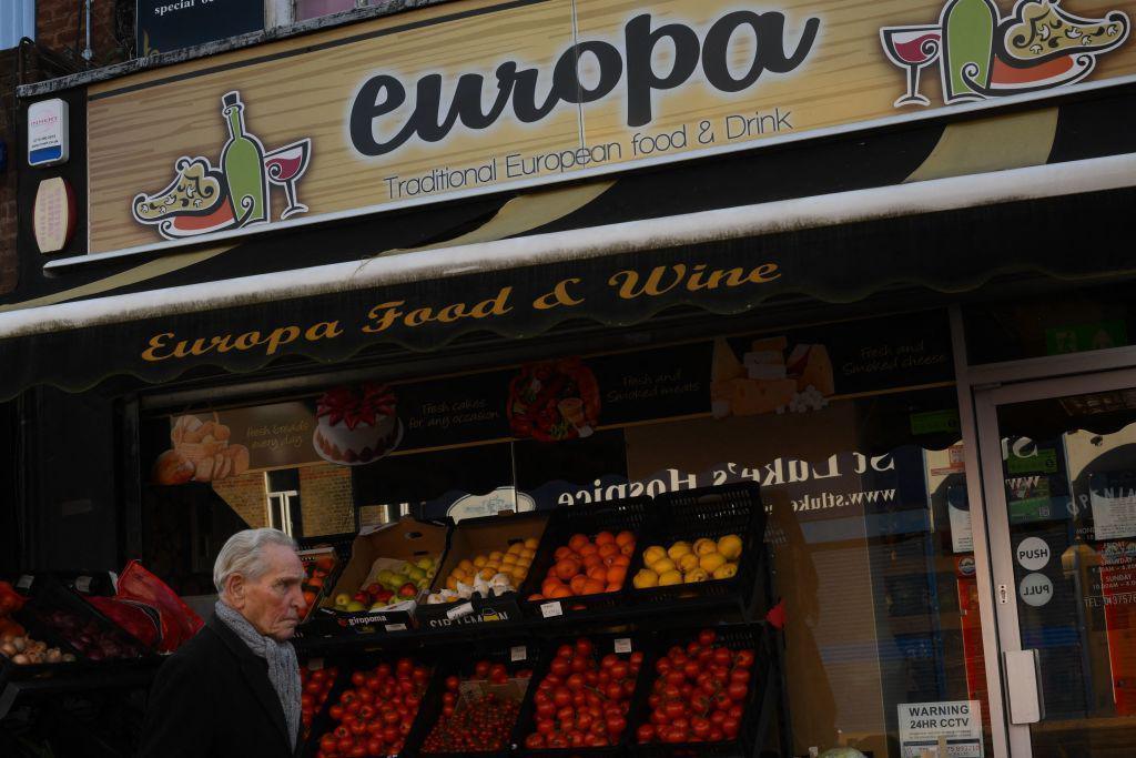 A man in a coat and with white hair walks past an eastern European grocery store in Grays, Essex, on January 18, 2023. There is fruit on display outside the shop, which is called "europa".