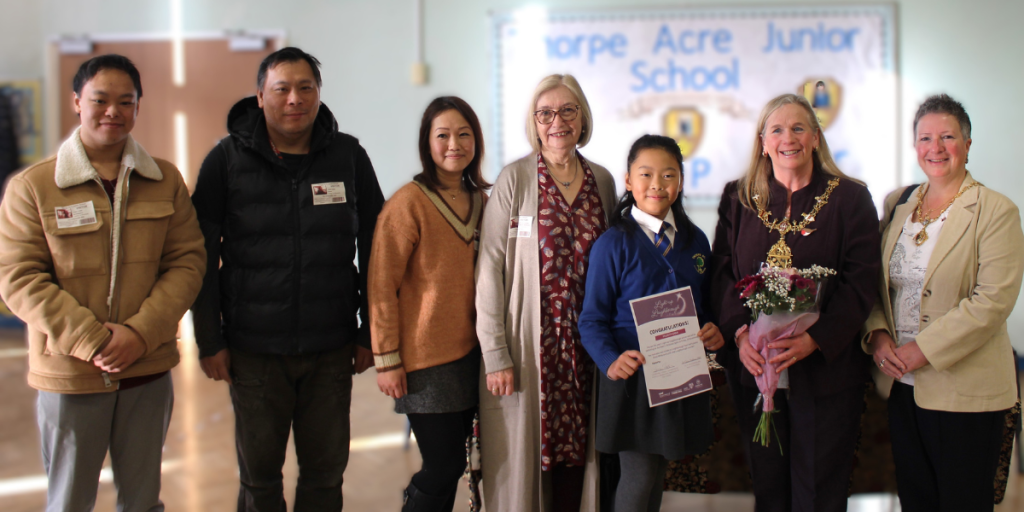 A row of people posing for a photo in a junior school hall alongside Felicity with a certificate, including smiling family members and the Mayor in her chain of office holding a bouquet of flowers