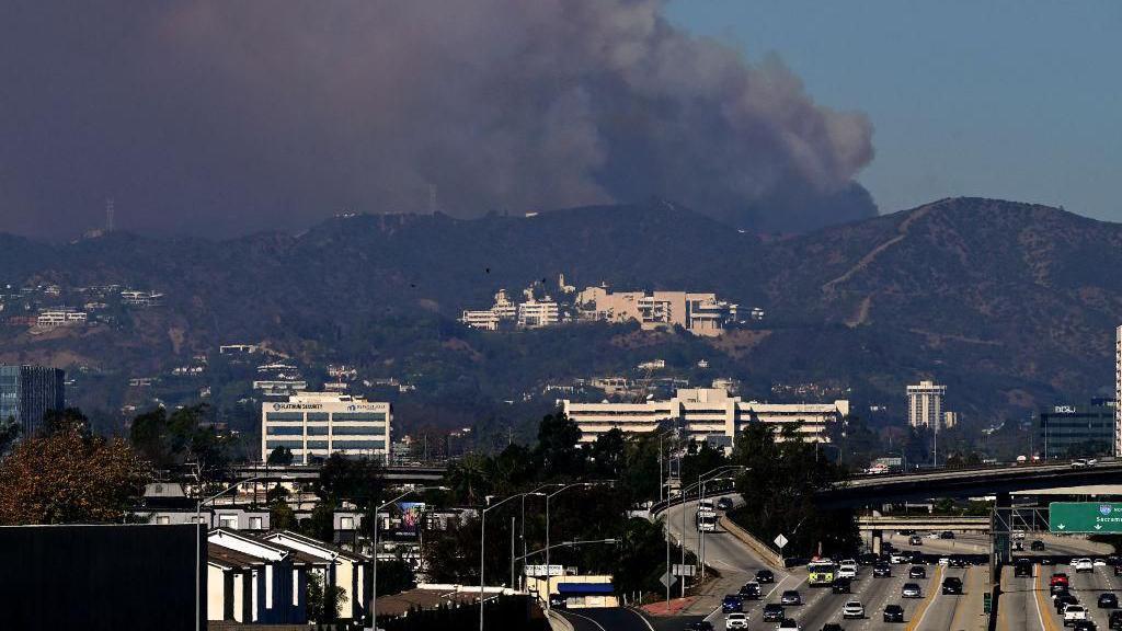 A long shot of Los Angeles with an enormous plume of smoke rising above hills on the edge of the city.