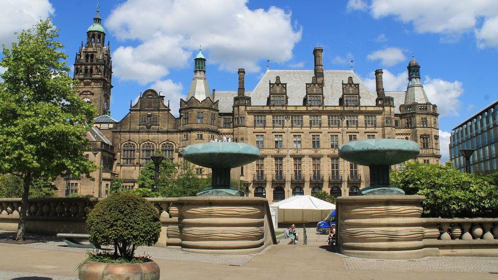 The outside of Sheffield Town Hall viewed from the entrance to the Peace Gardens, which features two fountains on either side of the staircase. The building was designed in a Gothic Victorian style with a clock tower, turrets and large arched windows.