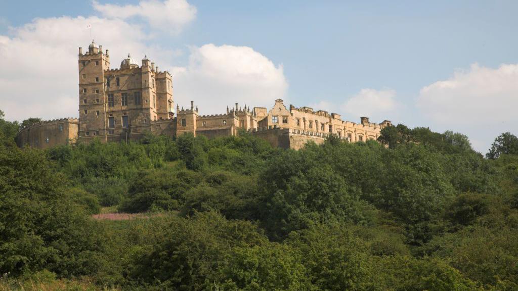 A view of Bolsover Castle - an ornate tower and associated ruined buildings - seen from slightly below looking up at its position on a steep wooded hill