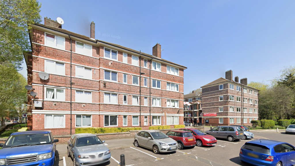 A four-storey red-brick block of flats with parked cars in front, under a clear blue sky.