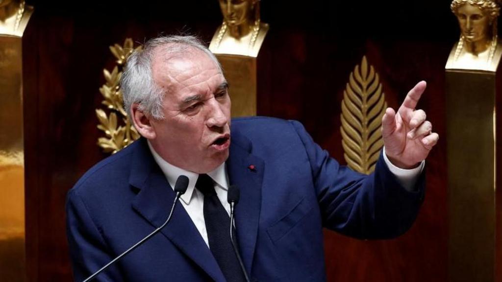 French Prime Minister Francois Bayrou speaks to parliament and points to the side. He wears a suit and speaks behind a podium. 