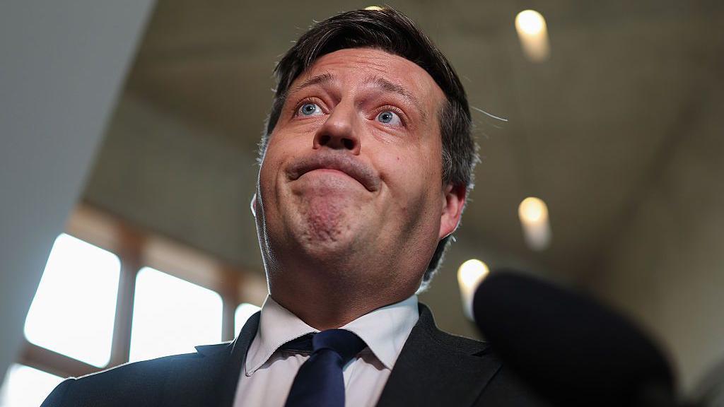 Jamie Hepburn is pulling a rueful face, standing in front of a microphone in the Holyrood building. He is wearing a dark suit, white shirt and plain dark blue tie. A set of high windows can be seen in the background.