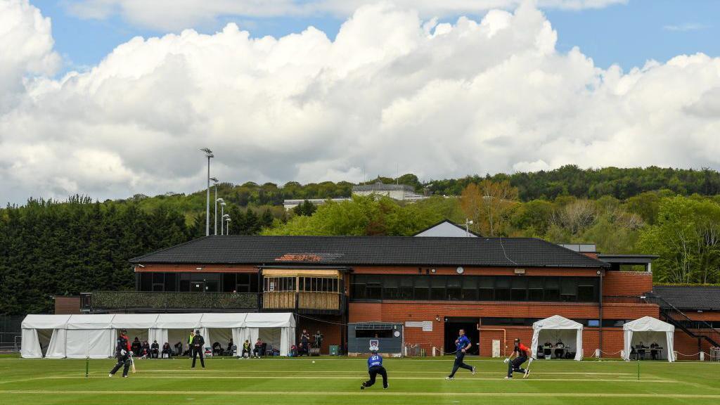 A large red brick building with a black roof. In front of it, a green cricket pitch with several people running.