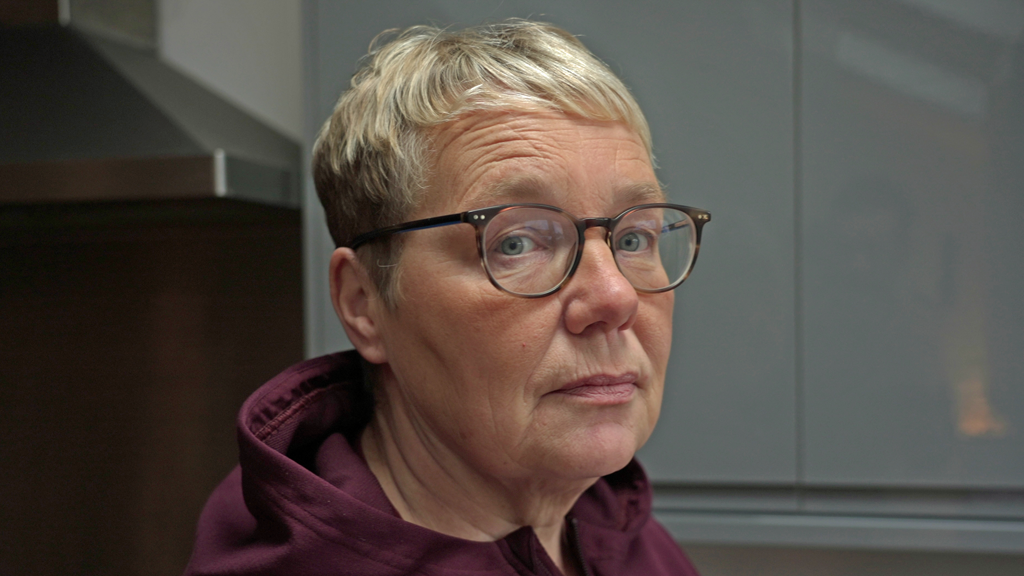 Headshot of Sue, who has short blonde hair and tortoiseshell framed glasses. She is wearing a burgandy hooded top and is pictured in a kitchen, with white cabinets behind her.