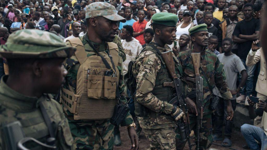 Armed M23 fighters in military uniform and green berets stand in front of a crowd of people. 