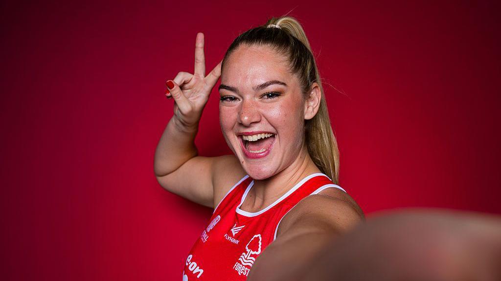 Rolene Streutker poses for a portrait. She has blonde hair, tied back, and is smiling into the camera as she makes a gesture with her right hand