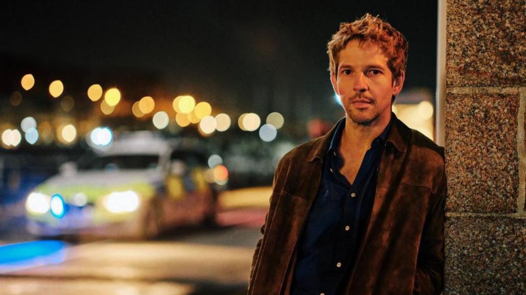 Actor Damien Molony in character as Jim Bergerac. He is leaning on a brick wall with a police car with its lights on blurred in the background. It is night time.