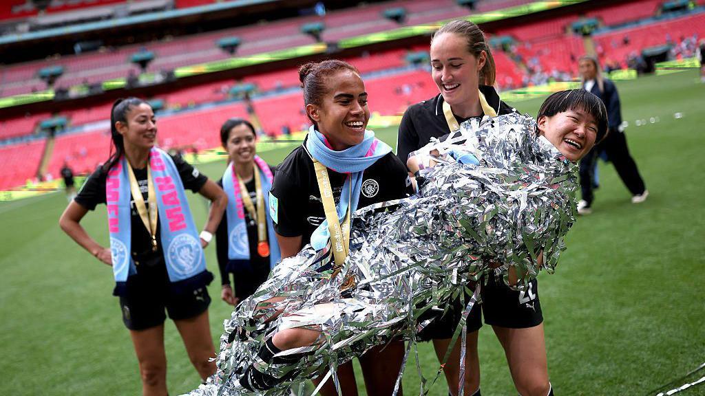 Mary Fowler and Sydney Lohmann carrying Aoba Fujino, wrapped up in silver ribbons, after the game.