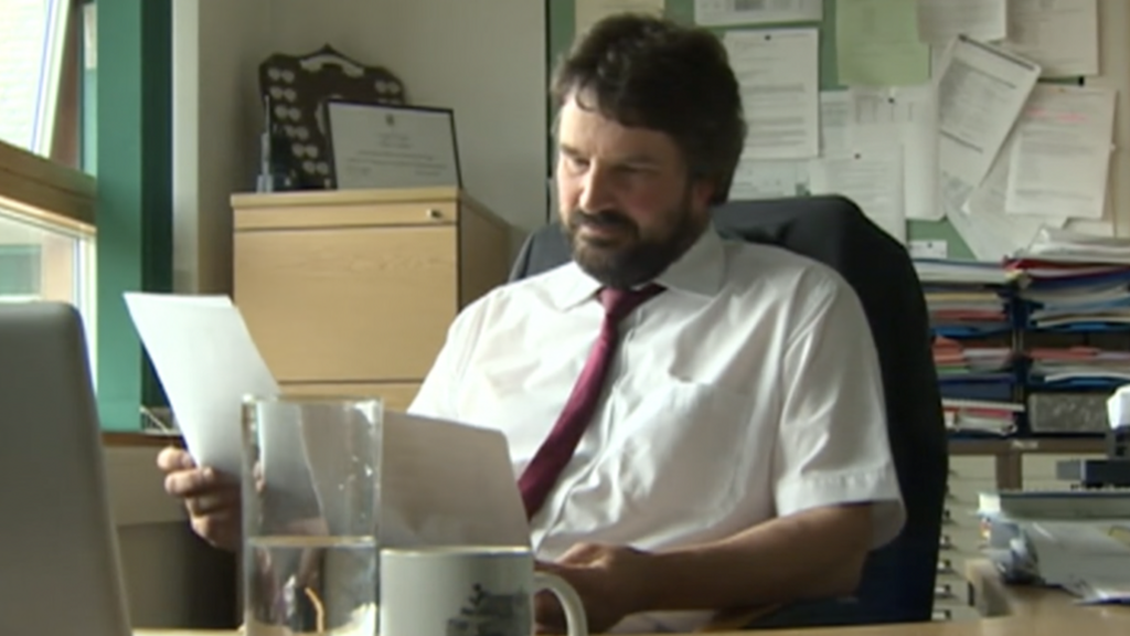 A man sat at a desk, looking at some paperwork. He's wearing a white shirt and red tie. A messy background full of books and paperwork. 