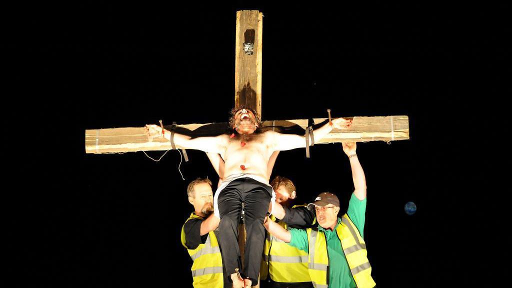 Michael Sheen topless tied to a crucifix, with three men in high vis jackets propping him up