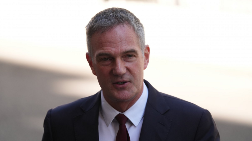 Business Secretary Peter Kyle mid-shot, he's wearing a dark suit, white shirt and dark red tie. He's walking outdoors, the background sunny but blurry