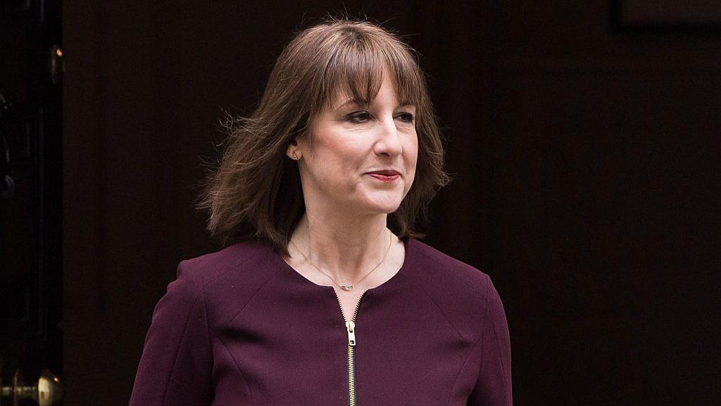 Rachel Reeves is dressed in an aubergine suit which zips up. She is pictured from the torso up and has brown mid length hair, with a fringe and wears a berry coloured lipstick. She has a small necklace and a neu.