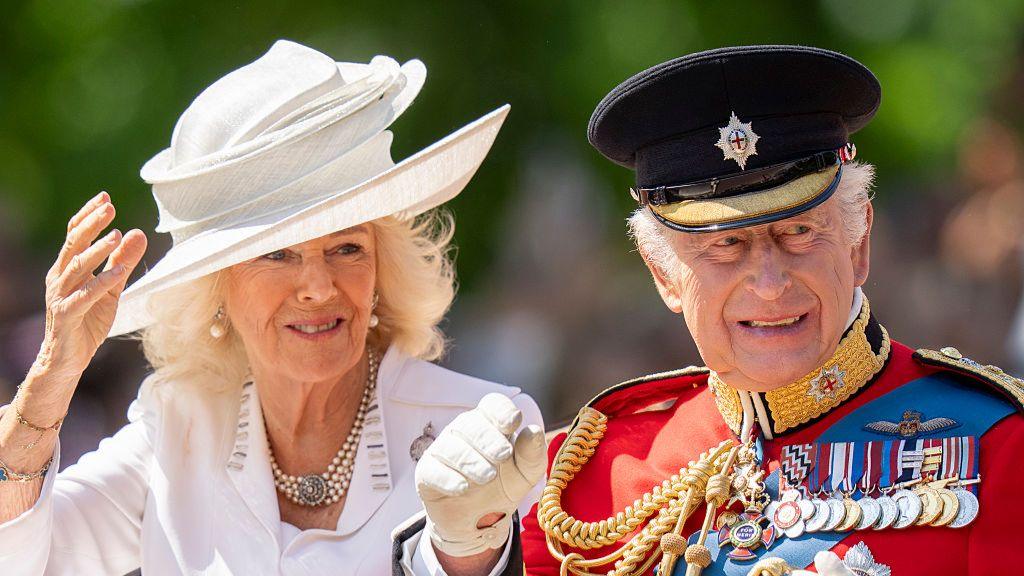 Charles, in a red uniform jacket and black hat, waves alongside Camilla, who wears a white jacket and hat. 