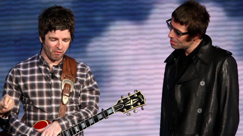 Noel Gallagher and Liam Gallagher "Che Tempo Che Fa" Italian TV Show on November 9, 2008 in Milan, Italy Noel looks down laughing holding a guitar. he is wearing a check shirt. Liam (right) looks at the presenter who is out of shot. he is wearing a leather jacket and sunglasses