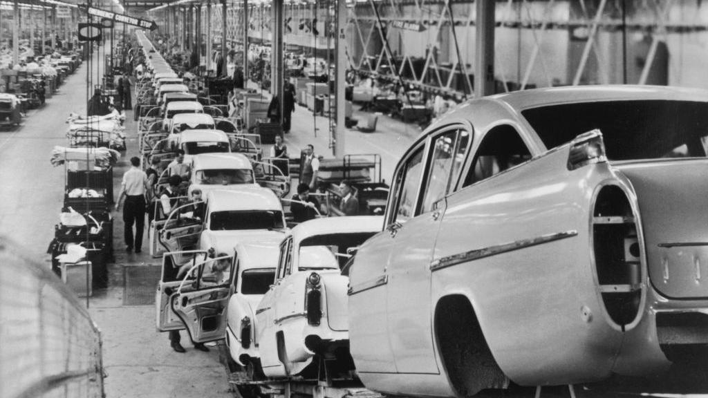 A black and white photo of a long car production line in a large warehouse. The cars in the foreground have no wheels and workers can be seen working on the vehicles