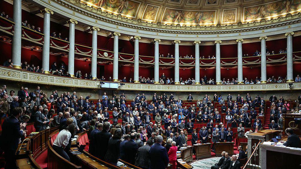 A wide shot inside the parliament building, you can see many people, all standing. The room is very beautiful, with intricate gold and pillars, and you can see the ornately painted ceiling.