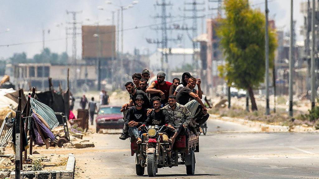 Men ride on a motorised cart along Salah al-Din road in Deir al-Balah, as smoke billows in the background, in the central Gaza Strip (21 July 2025)