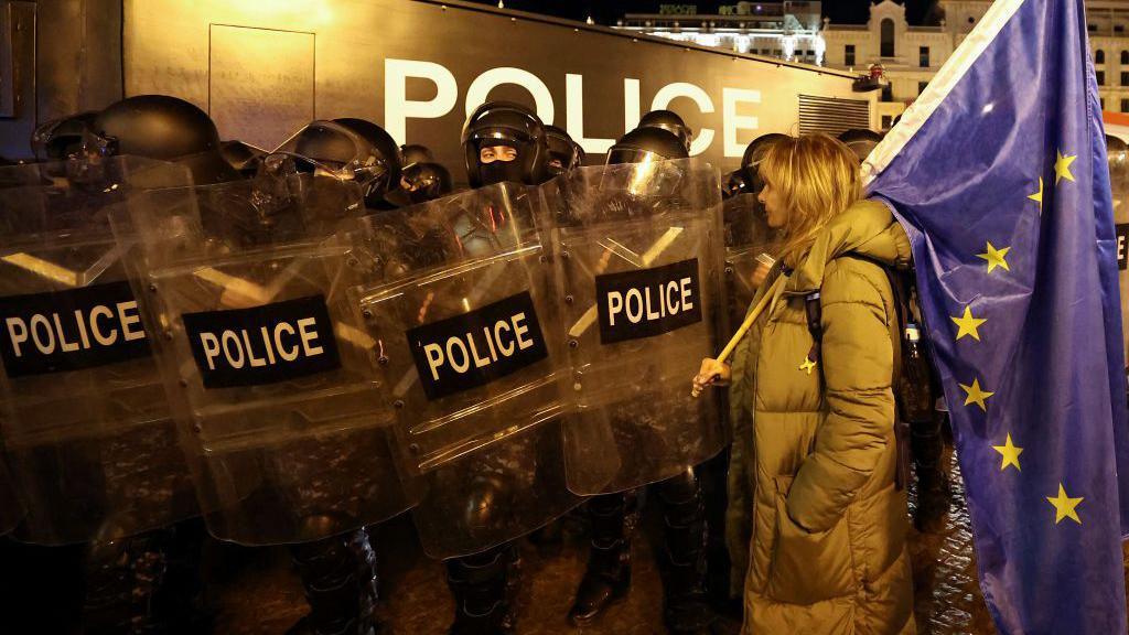 A protester in Tbilisi, Georgia stands in front of a line of riot police holding a large European Union flag.  